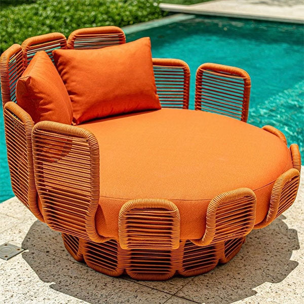 A round outdoor lounge Daybed with a bright orange cushioned seat and backrest, surrounded by a woven wicker-style base, shown beside a swimming pool on a stone patio.