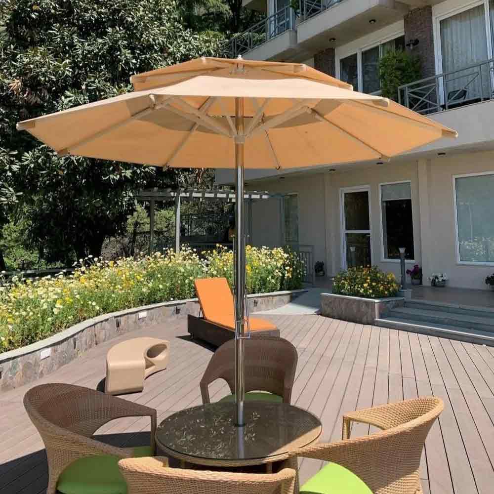 A large beige patio umbrella centered over a round outdoor dining table with wicker chairs on a wooden deck, with a modern house and landscaped garden in the background.