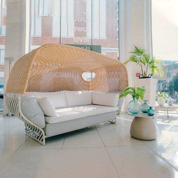 A modern outdoor lounge daybed with a white woven frame and a large arched natural rattan canopy, styled in a bright, contemporary patio lounge setting.