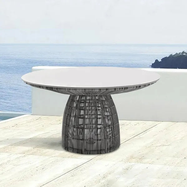 A round outdoor dining table with a light gray top and a dark woven pedestal base, set on a stone patio with a white wall and ocean view in the background.