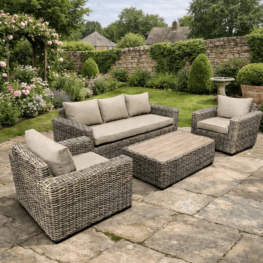A 4-piece outdoor patio furniture set made of woven wicker with light beige cushions, including a sofa, two armchairs, and a rectangular coffee table, arranged on a stone patio in a lush garden setting.