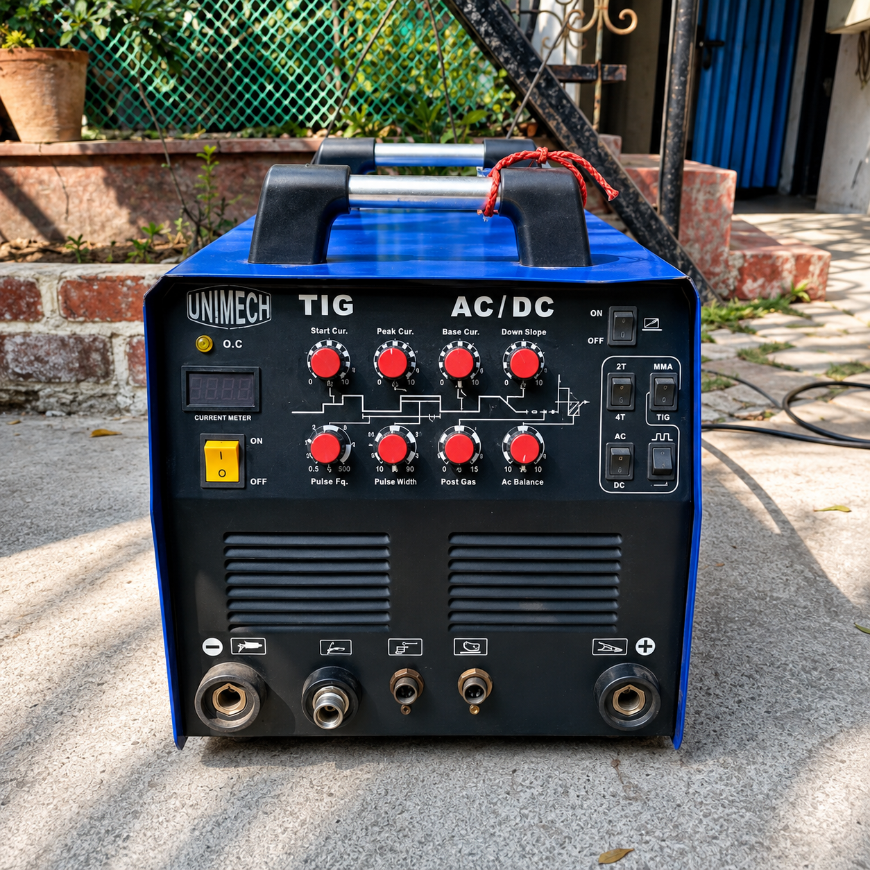 A blue and black UNIMECH TIG / AC/DC welding machine photographed outdoors on a concrete surface with a brick wall, fence, and gate in the background.
