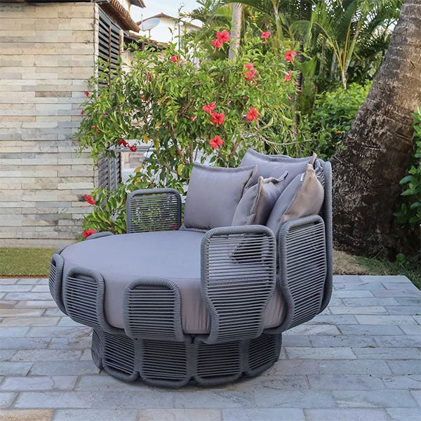 A round outdoor daybed with a gray Rope frame and matching gray cushions, set on a stone patio with tropical plants and flowers in the background.