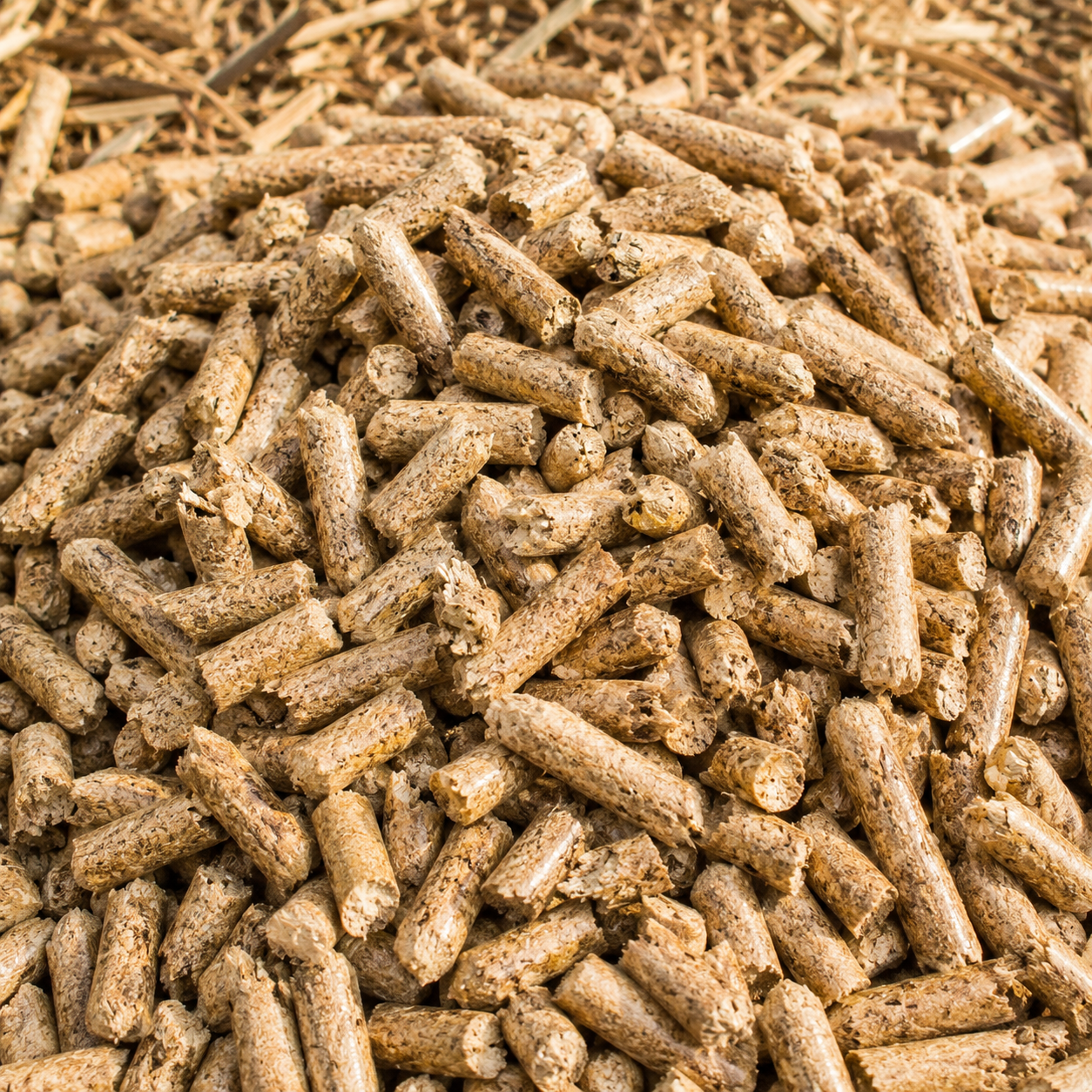 A close-up pile of brown cylindrical pellet pieces, likely wood or agricultural biomass pellets, filling the frame against a similar straw-like background.