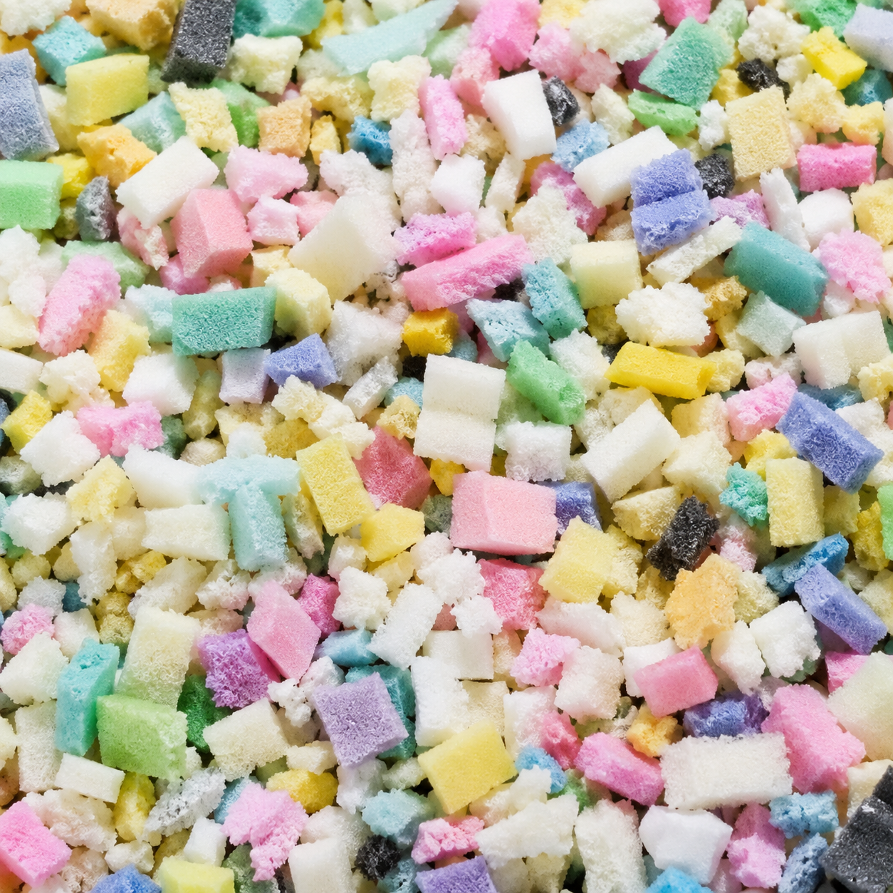 A close-up, top-down view of a colorful candy or confection mix consisting of many small, irregularly shaped sugar cubes in pastel shades of pink, yellow, green, blue, purple, and white.