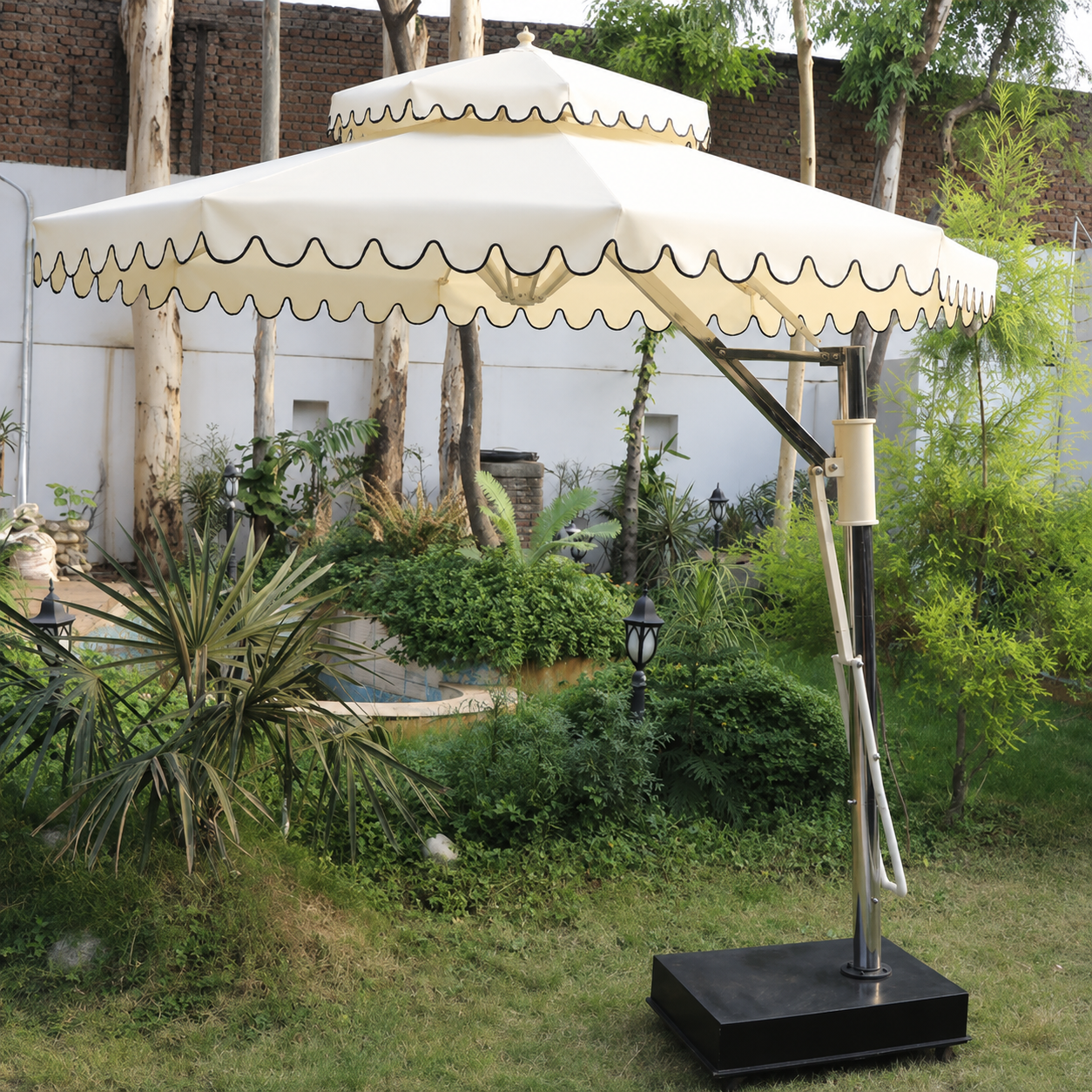 A large outdoor patio umbrella with a cream-colored canopy and scalloped trim, mounted on a black metal frame with a square base, set in a garden environment.