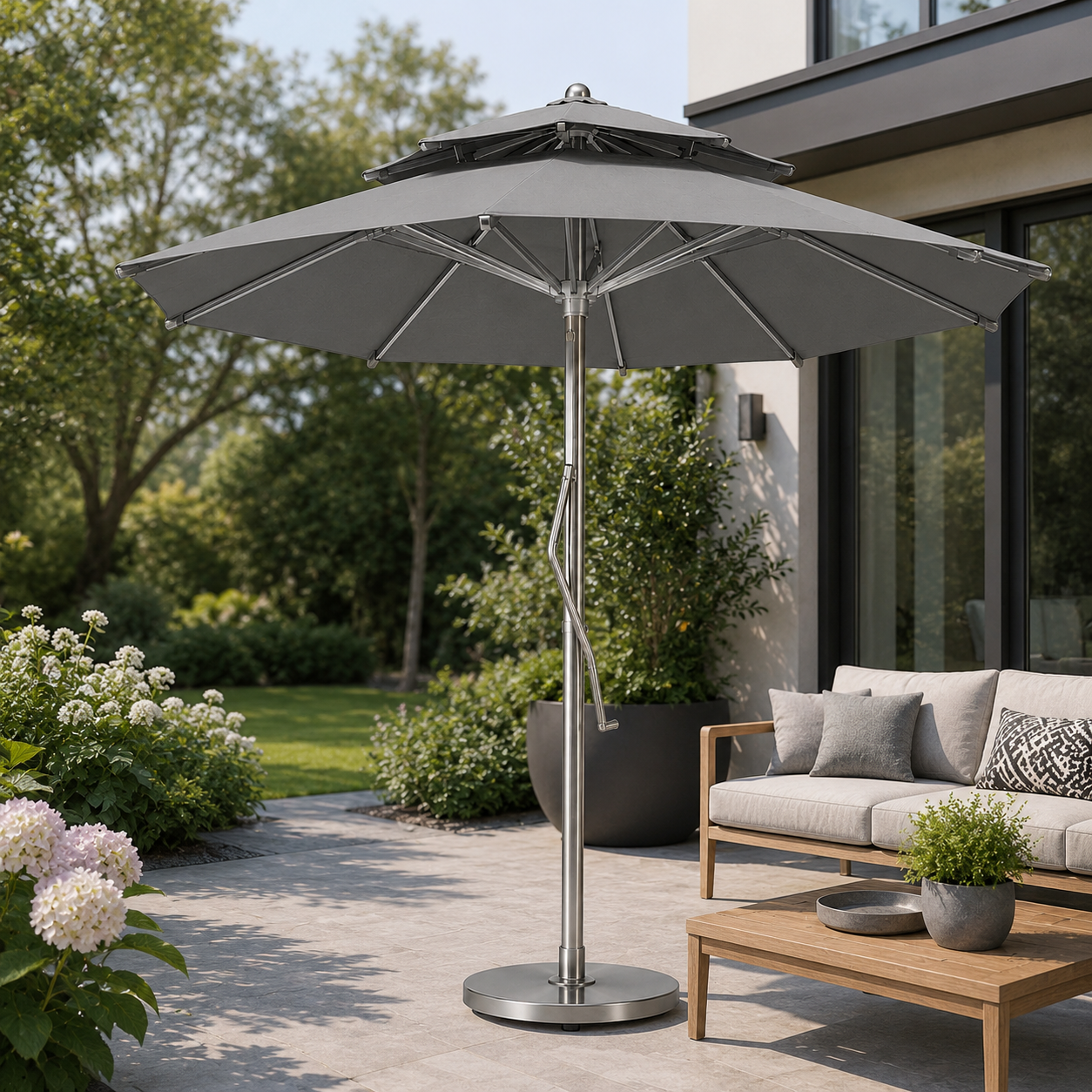 A large gray patio umbrella with a double-tier canopy and a silver metal pole stands on a stone patio next to a modern outdoor sofa with cushions.