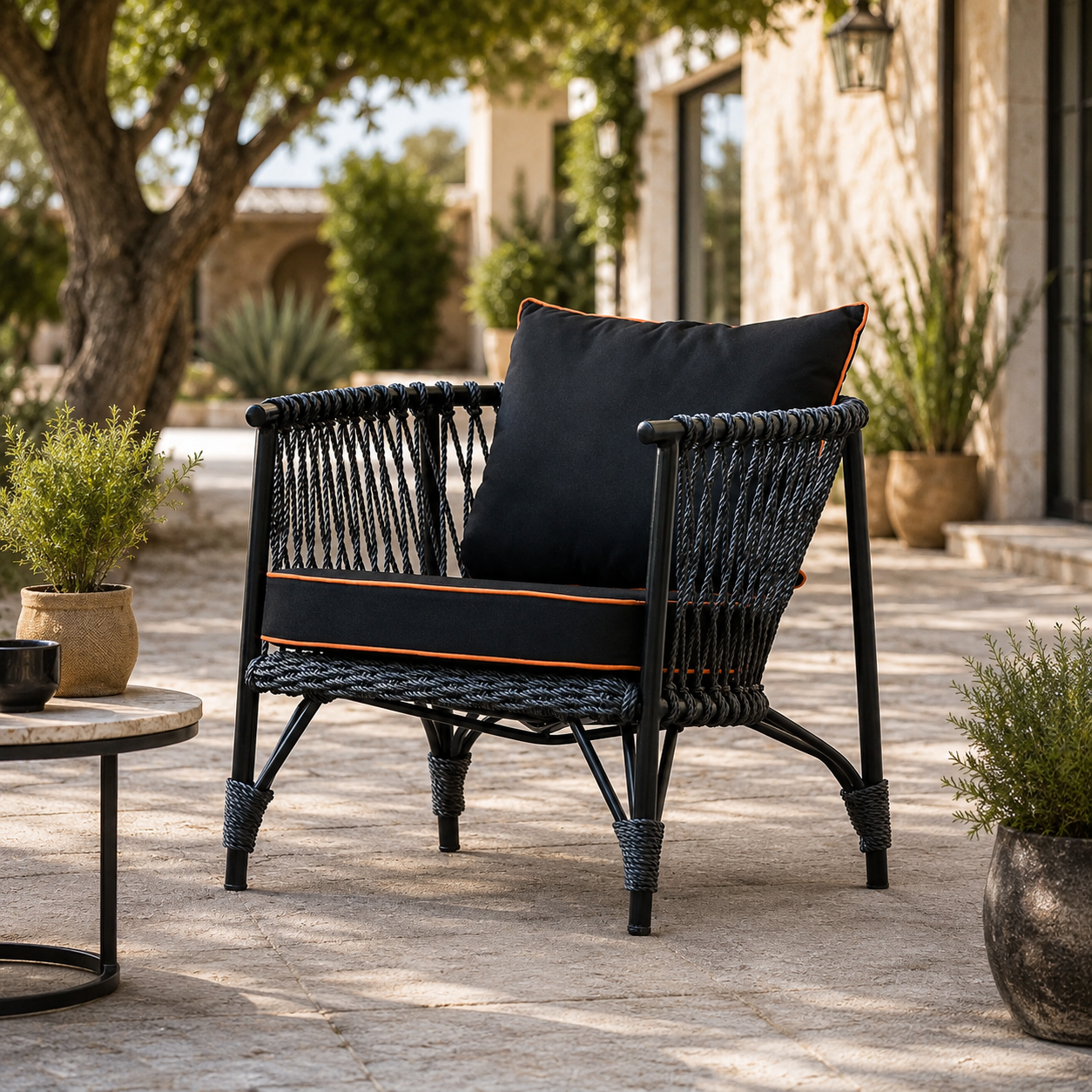 A black woven outdoor armchair with black cushions and orange piping trim, placed on a stone patio in a garden setting.