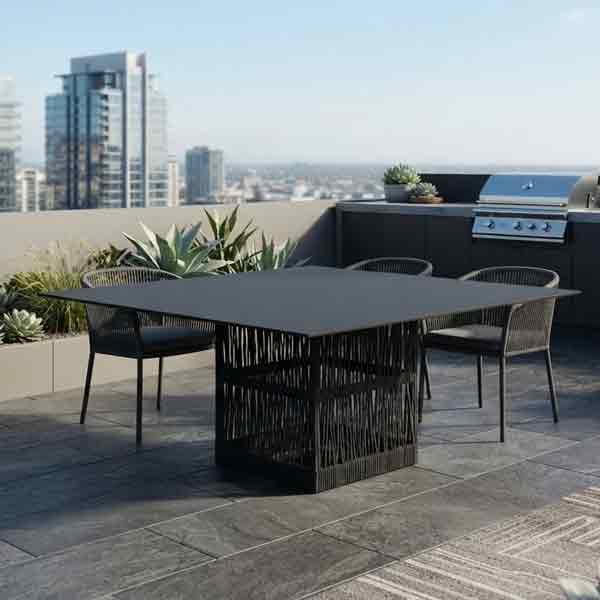 A modern outdoor dining set on a rooftop terrace, featuring a rectangular black table with a woven base and four matching woven chairs, with a stainless steel grill and city skyline in the background.