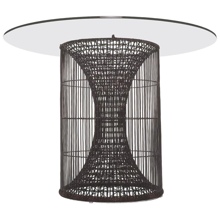 A top-down view of a round, woven dark gray wicker or rattan planter or decorative base placed on a light gray circular surface over beige stone tiles.