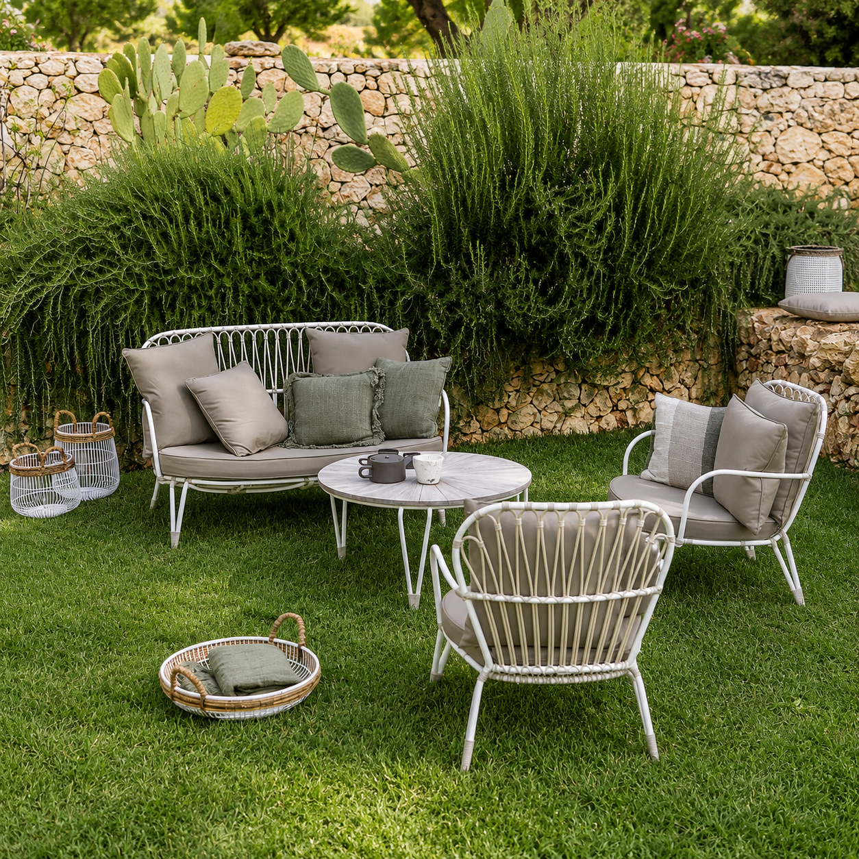 Outdoor patio furniture set on a green lawn with a stone wall and plants in the background. The set includes a white metal sofa with beige cushions, two matching armchairs with gray cushions, a round white coffee table, and a woven tray with a cushion.