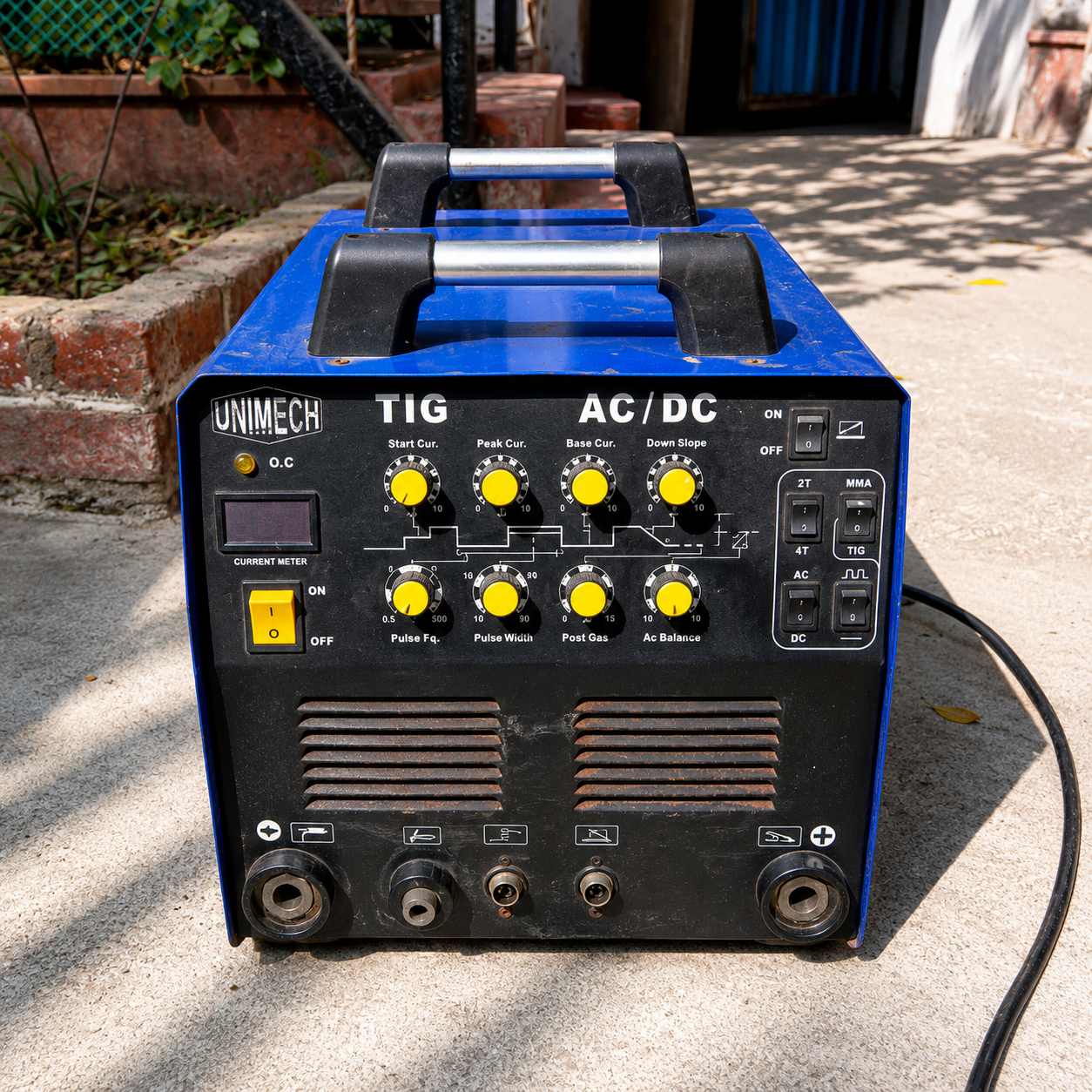 A blue and black UNIMECH TIG AC/DC welding machine sitting outdoors on a concrete surface with its power cord visible.